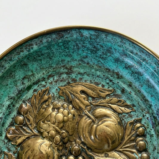 Close-up macro detail of the front surface of an oxidized Italian brass plate, showcasing the contrast between the raised, polished gold-toned brass grape leaves and the deep, mottled turquoise and teal verdigris acid-etched patina.