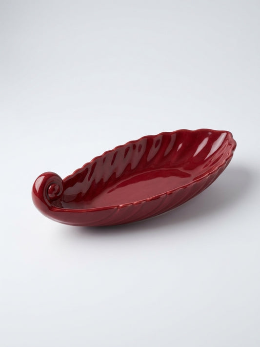 High-angle shot of a deep burgundy ceramic leaf-shaped dish with a scrolled stem end and scalloped edges, resting on a white surface.