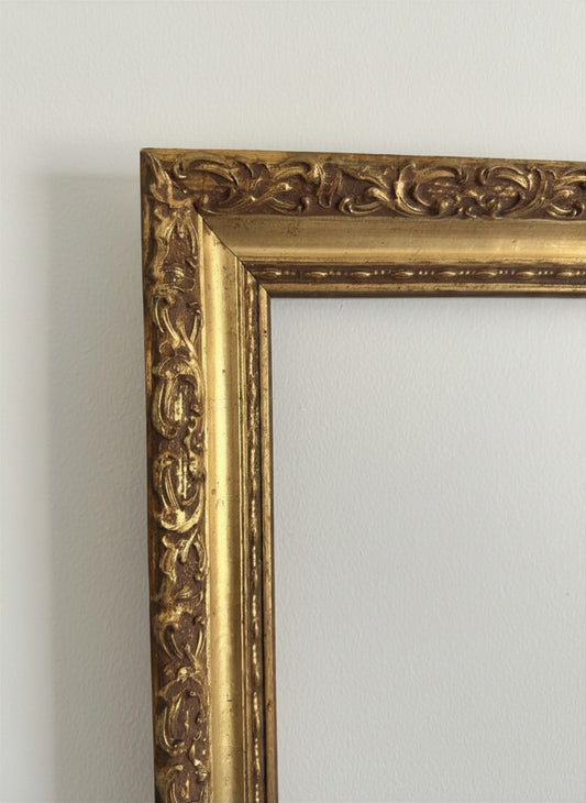 Micro detail close-up of a vintage gilded gold frame highlighting the dimensional, hand-carved style scroll pattern and original aged patina. Shows dark undertones and a textured, warm metallic finish. Perfect for styling modern abstract art contrasts or classic family portraits.