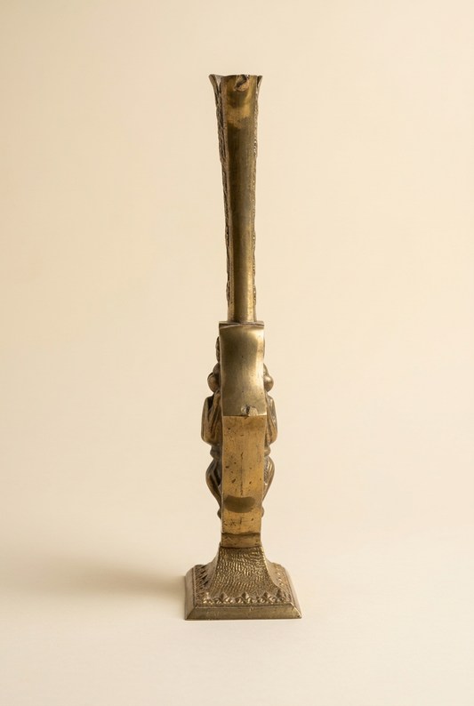 Side profile of an ornate brass vessel showing its slender neck, projecting floral relief, aged patina, and rectangular pedestal base.