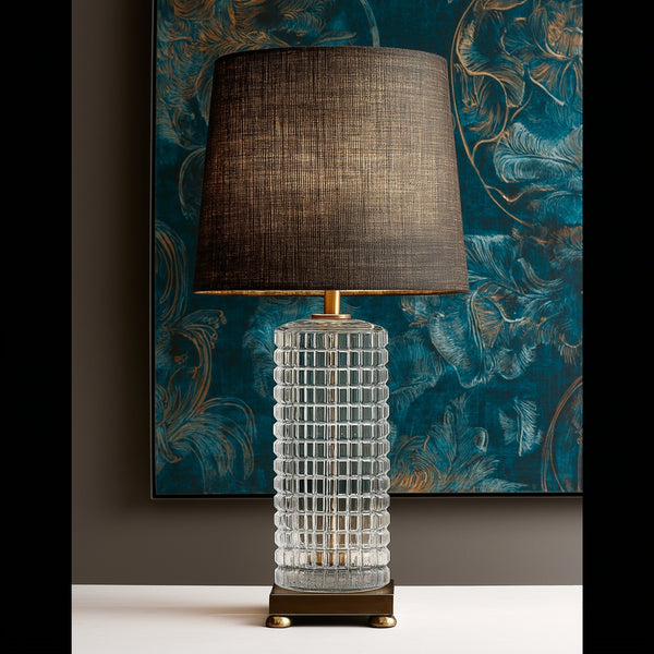 A sculptural table lamp featuring a textured, gridded clear glass cylinder base and a dark woven fabric drum shade, styled against a moody dark academia backdrop with teal and gold abstract wall art.