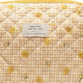 A quilted cosmetic pouch with a beige and white gingham pattern, decorated with small yellow floral motifs. It features a top zipper with a gold pull and a small white label on the front.