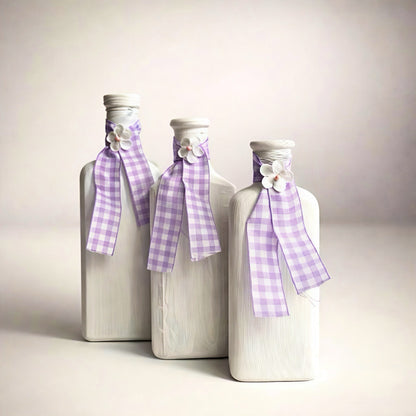 Set of three decorative white glass bottles with lavender checkered ribbons and floral accents.