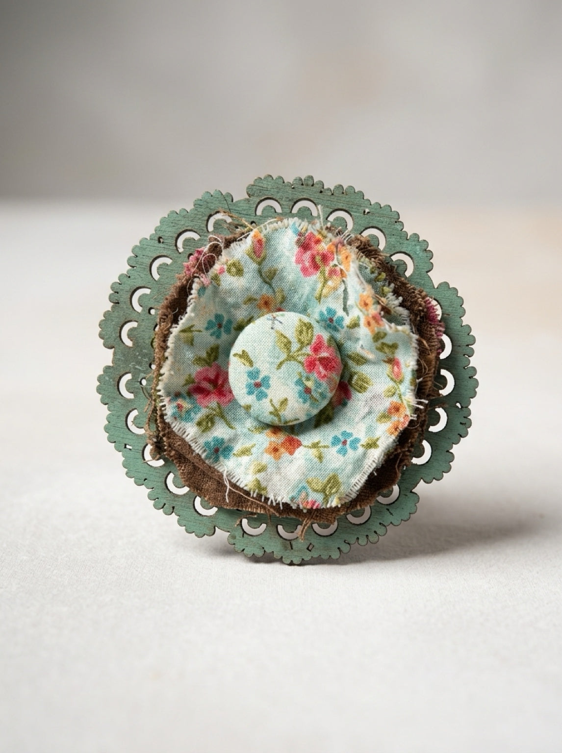  Close-up of floral fabric rosette with a covered center button, frayed edges, and scalloped green backing.