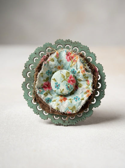  Close-up of floral fabric rosette with a covered center button, frayed edges, and scalloped green backing.