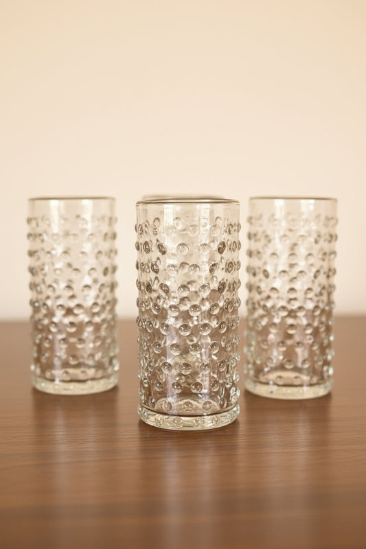 Set of 2 clear cylinder highball glasses with raised hobnail beaded texture sitting on a dark wooden tabletop, 6 inches tall. Arranged on wooden surface 