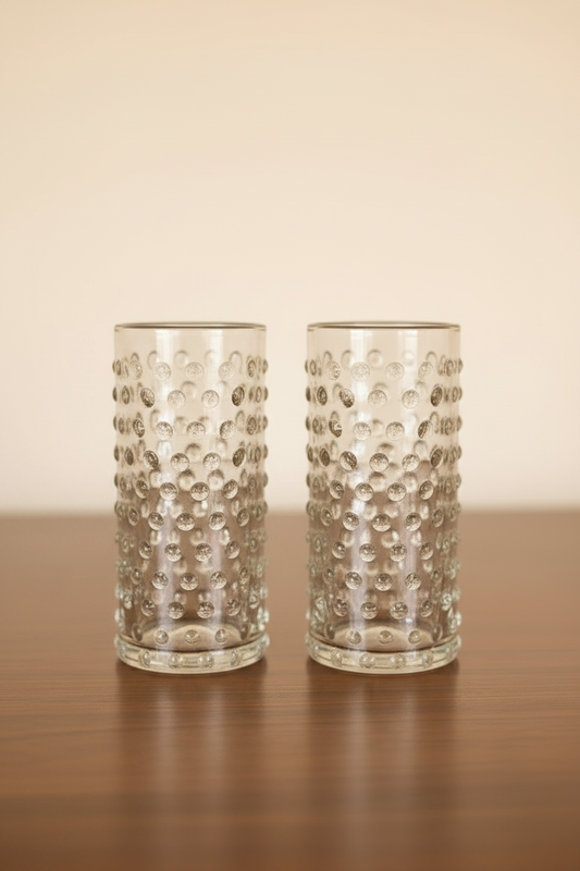 Set of 2 clear cylinder highball glasses with raised hobnail beaded texture sitting on a dark wooden tabletop, 6 inches tall.