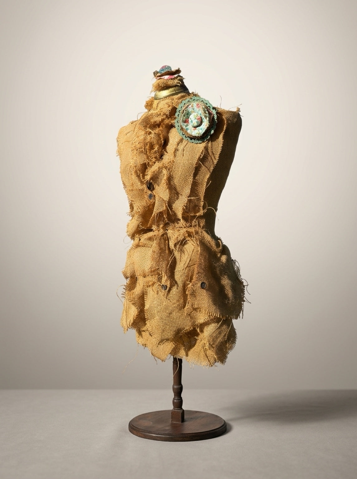 Angled view of burlap dress form sculpture showing floral shoulder rosette, button accents, and layered fabric skirt.