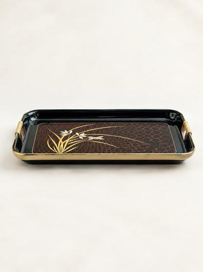 Low front view of a black lacquer floral serving tray showing raised sides, gold-tone rim, and cane-wrapped cut-out handles.
