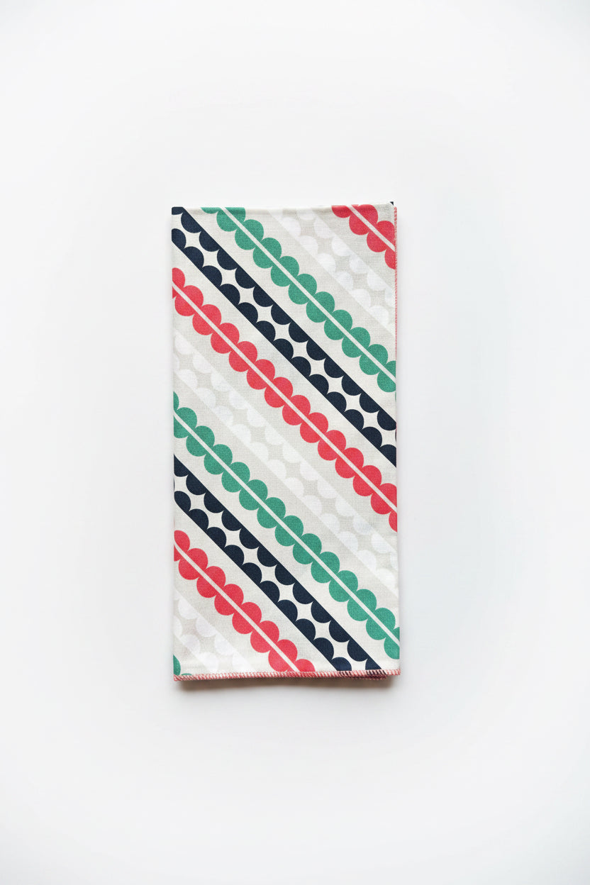 Single Matilda cloth napkin folded lengthwise displaying hand-printed diagonal dot-and-stripe geometric pattern in coral, teal, and navy on crisp white cotton