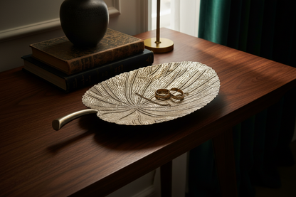 A high-end lifestyle interior design photograph of a cast metal elephant ear leaf catchall tray. The tray features a highly detailed, stippled, and deeply ribbed organic texture with a distinct raised, smooth polished stem handle. The finish is a luxurious, low-luster champagne gold with soft silver-gilt highlights that subtly catch the ambient light.

The leaf tray is positioned as a striking centerpiece on a dark, rich walnut wood console table. It is styled intentionally as an eclectic catchall, holding 