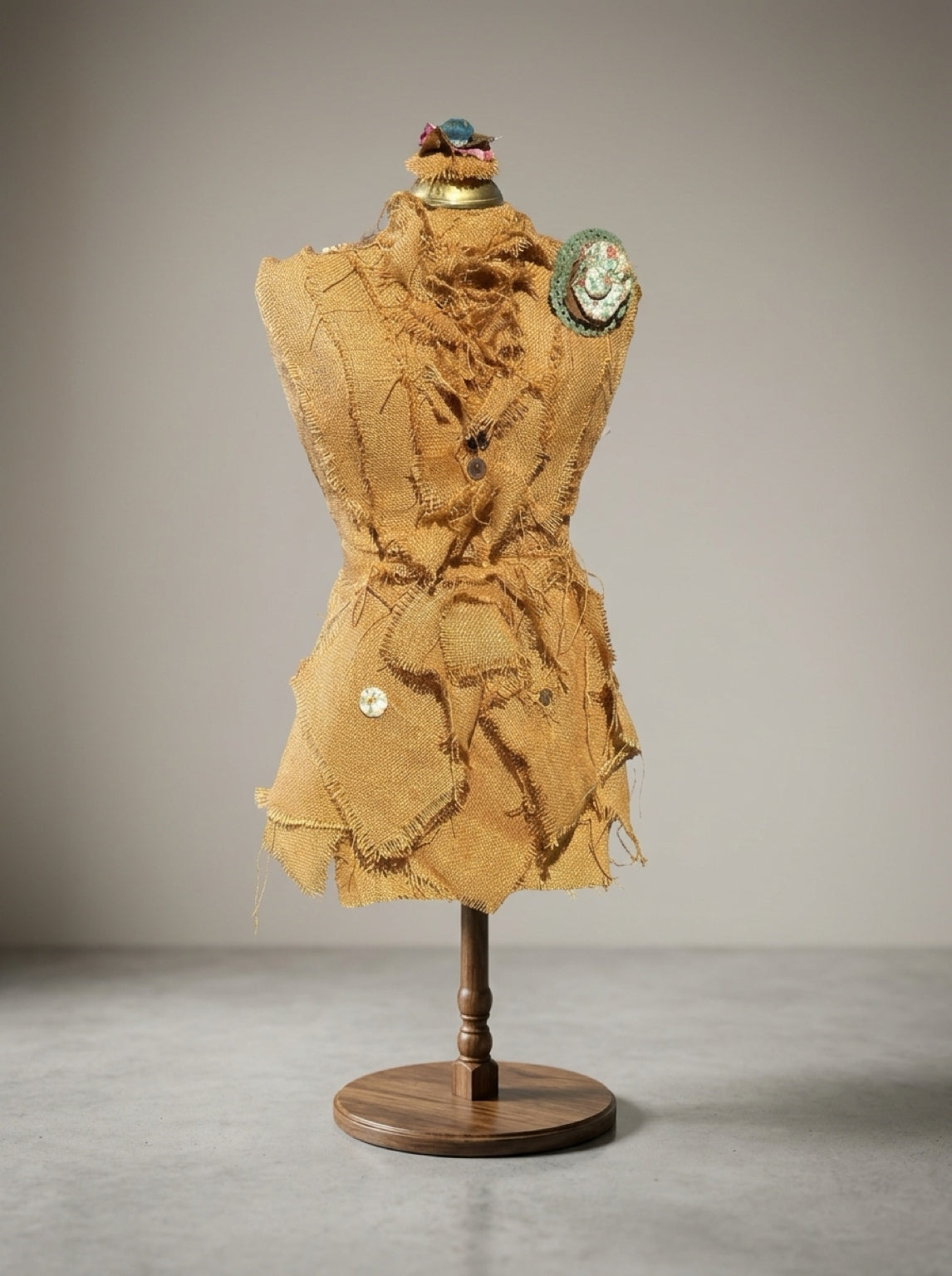  Handcrafted burlap dress form sculpture with frayed fabric layers, floral shoulder rosette, and round wooden pedestal.