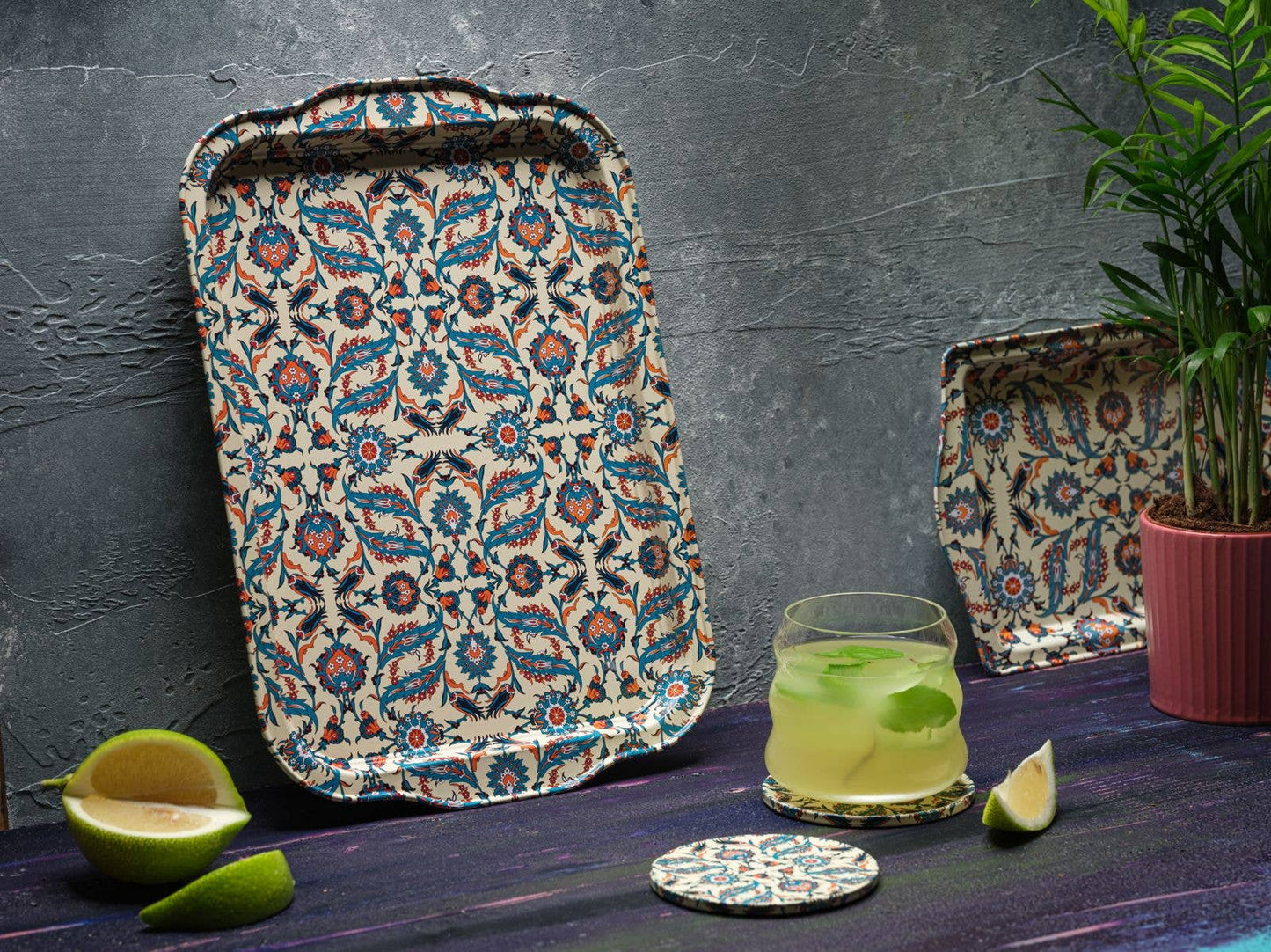 Beste patterned tin serving tray by The Marian Collection styled upright against a textured dark gray wall. This rectangular platter with a vibrant teal and red Turkish motif is paired with a matching coaster, a fresh mint cocktail, lime slices, and a small potted palm, showcasing a moody maximalist bar setup.