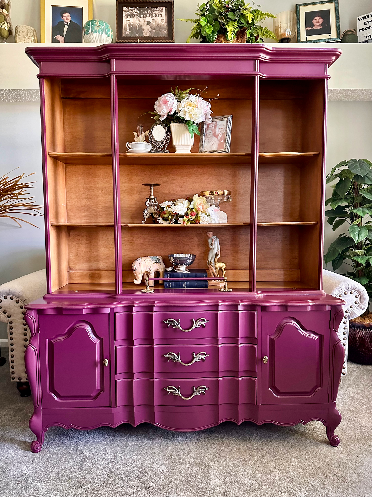 Vintage wooden hutch painted in a bold magenta finish with natural wood shelves, styled with eclectic décor including framed photos, florals, silver accents, and small figurines in a cozy living room setting.