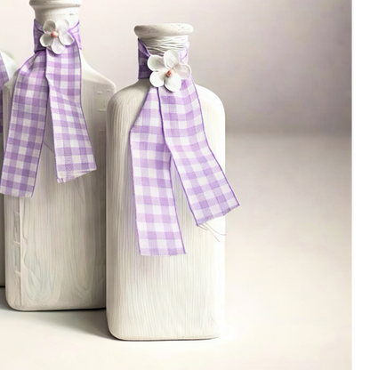 Set of three decorative white bottles with purple checkered ribbons and floral accents for home decor.