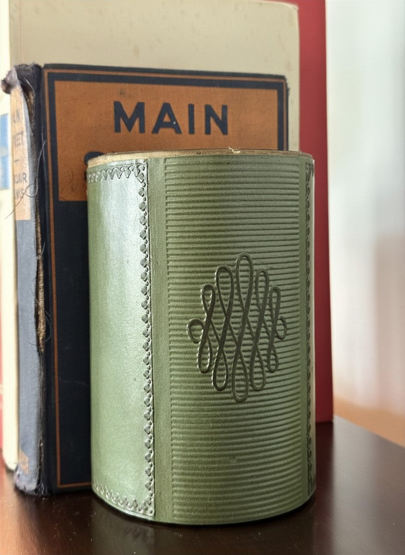 Close-up of vintage mid-century sage green leather half-moon bookends showing detailed stamped scalloped border and horizontally ribbed leather texture resting against a worn antique hardcover copy of Main Street by Sinclair Lewis.
