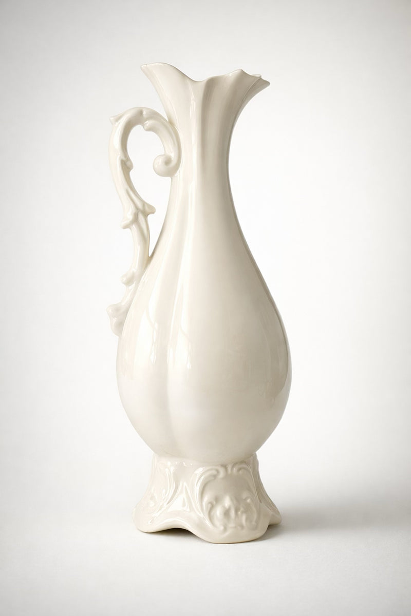 Cream-colored vintage ceramic pitcher with a tall tapered body, scalloped wavy rim, ornate curved handle, and embossed base detailing, styled against a clean neutral background.