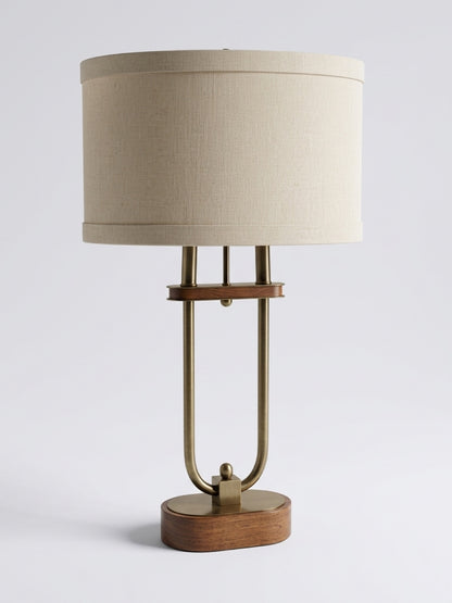 Mid-century modern table lamp featuring a antique brass u-shaped frame, stacked dark walnut wood oval base, and beige textured linen drum shade set against a plain white studio backdrop.