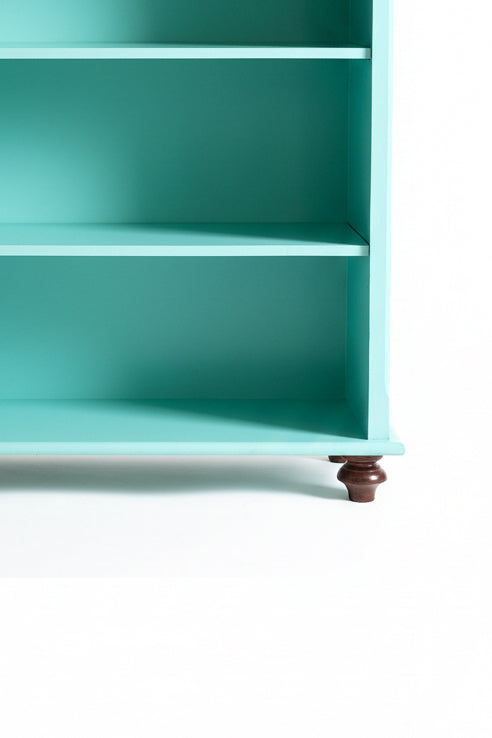 Detailed close-up view of the lower shelves and base of a refinished solid wood bookshelf. The vibrant teal painted surface features a smooth satin poly-acrylic topcoat over clean wooden shelving. The unit is supported by a beautifully turned dark wood bun foot in a rich espresso finish, highlighting the structural elevation and artisan quality of the furniture piece.