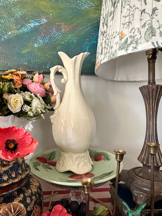 Glossy cream ceramic pitcher with a flower-like flared rim, decorative scroll handle, and sculpted embossed base, photographed in soft lighting to highlight its craftsmanship.