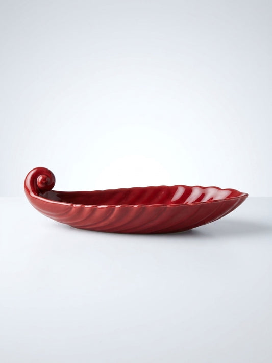 Side profile view of a deep red glazed ceramic boat-shaped vanity dish featuring a scrolled end and ribbed texture on a white background.