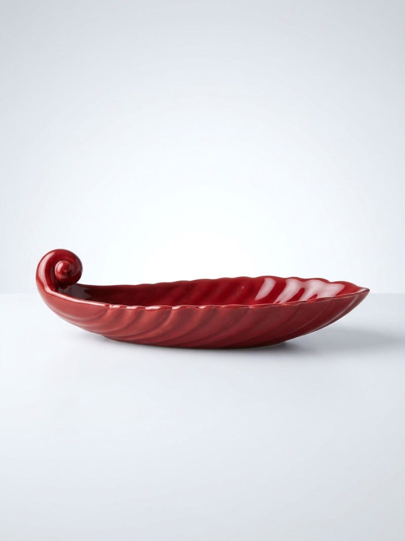 Side profile view of a deep red glazed ceramic boat-shaped vanity dish featuring a scrolled end and ribbed texture on a white background.