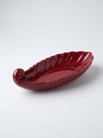 High-angle shot of a deep burgundy ceramic leaf-shaped dish with a scrolled stem end and scalloped edges, resting on a white surface.