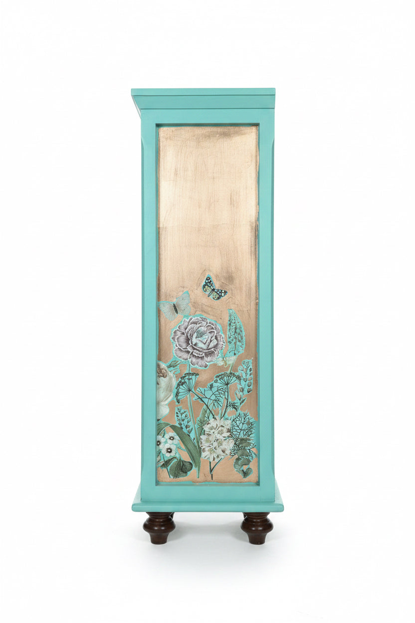 Left side recessed panel view of the custom refinished teal bookcase. The background is a hand-applied metallic copper leaf foil that reflects light beautifully. Layered over the copper is a detailed botanical decal featuring a gray and white peony flower surrounded by wild botanicals and realistic fluttering butterflies, showcasing the signature Florentine-inspired style of The Marian Collection.