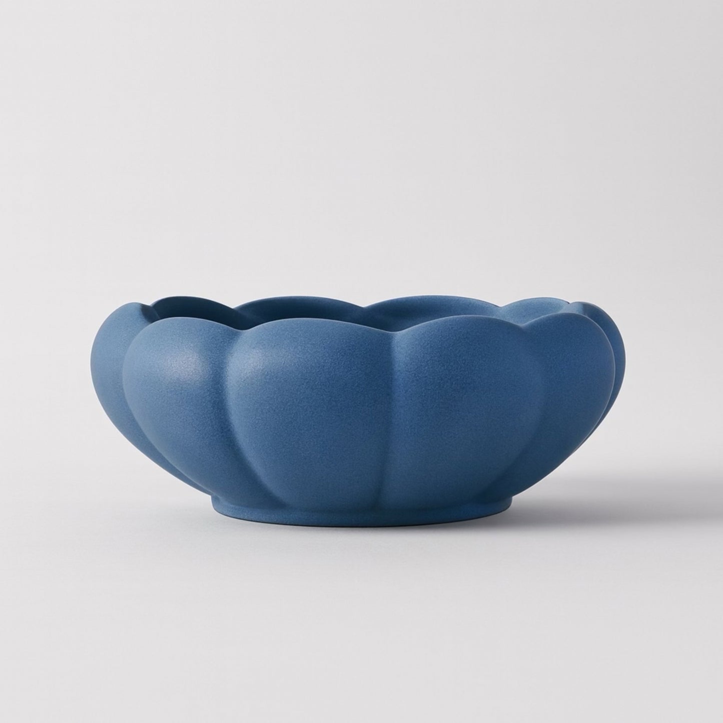 Matte blue ceramic bowl with scalloped, petal-like edges resembling a flower or pumpkin. Photographed against a white background to highlight its sculptural form and soft finish.