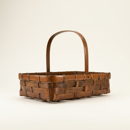 Side profile view of a shallow rectangular vintage bamboo market basket featuring a central overhead carrying handle and a dark chestnut brown hand-woven design.