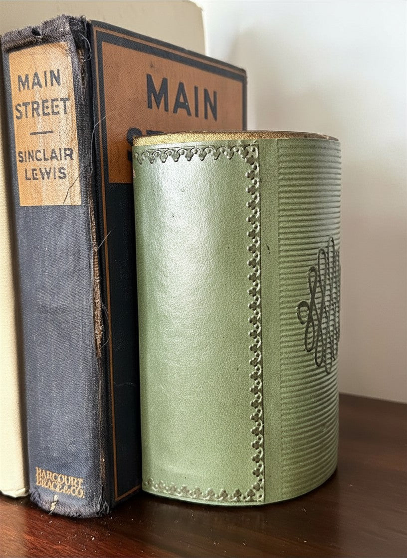 Side view detail of a mid-century olive green embossed faux-leather bookend highlighting a central looped scrollwork monogram emblem and horizontal ribbed banding on a wooden shelf next to a distressed dark blue book spine.