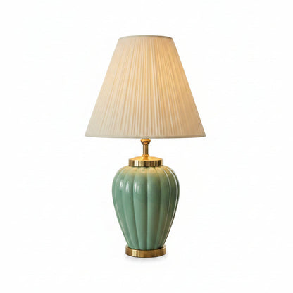 Teal lamp with a pleated beige shade on a table next to books and a picture frame, with a painting in the background.