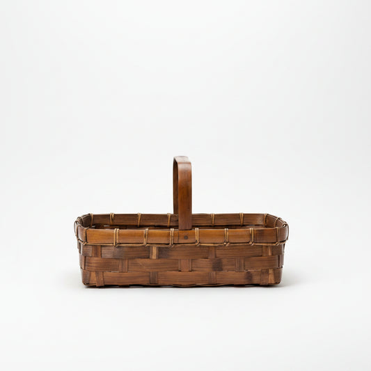 Three-quarter perspective of a vintage woven split bamboo gathering basket with a tall arched handle, tight cane bindings on the rim, and a rich dark amber brown patina, isolated on a clean cream background.