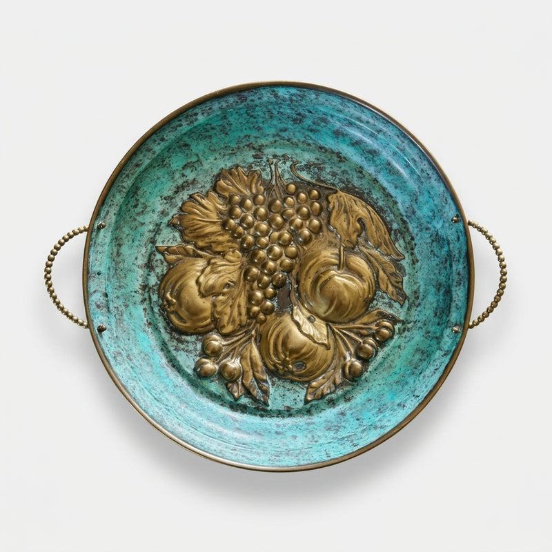 Top-down studio shot of a vintage Italian round brass tray on a white background, featuring a heavy-relief embossed fruit motif of grapes and peaches centered on a striking, textured turquoise-green verdigris patina with delicate beaded brass side handles.