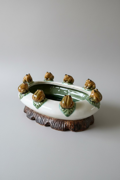 Vintage majolica style oval ceramic planter with eight molded frogs on green lily pads around the rim, white body with a brown faux-bois tree stump base.⁠
