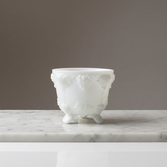Vintage white milk glass footed cachepot with an embossed grapevine pattern, sitting on a white marble slab against a light neutral wall.