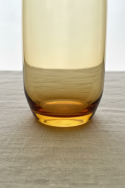 Close-up base detail of vintage amber glass highball tumbler showing thick heavy weighted bottom with warm golden-amber color concentration and smooth rounded base profile on linen surface
