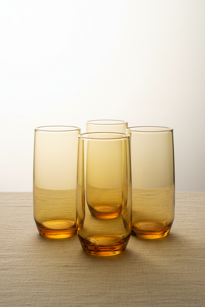 Set of four mid-century vintage amber glass highball tumblers grouped together on neutral linen surface showing warm honey-amber color, straight-sided cylindrical silhouette, and thick weighted bases
