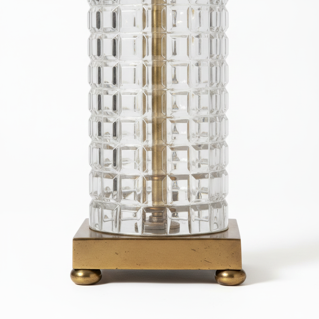 Close-up of vintage table lamp square antique brass plinth with four round ball feet, base of thick clear geometric waffle-grid glass column, light aged brass patina, white background