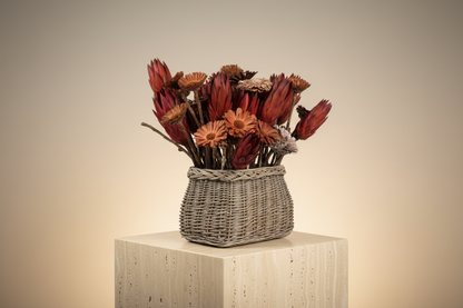 Dried Protea repens and solar wood bloom basket arrangement on a cream plaster pedestal in warm ambient light, three-quarter view showing full sculptural form of burgundy cone heads and amber daisy blooms in grey-washed woven reed basket