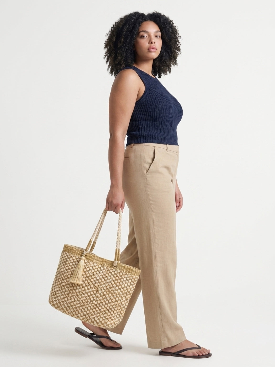 Side view of a model carrying the gold and cream checkerboard woven tote by its braided shoulder handles, paired with casual linen trousers.