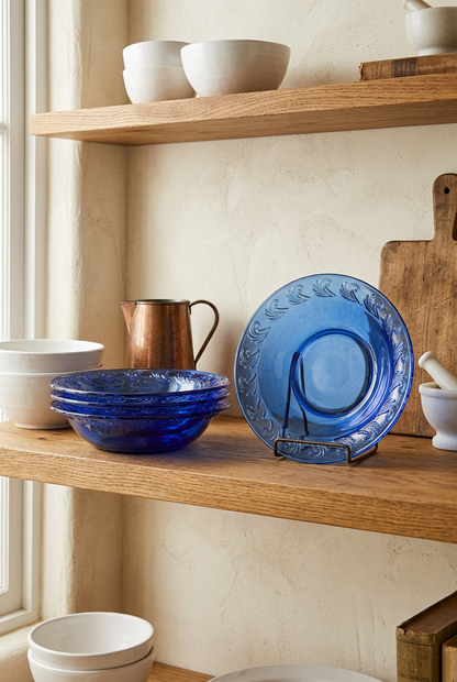 Cobalt blue glass pie dishes stacked and displayed upright on a wooden kitchen shelf beside a copper pitcher and white ceramics.
