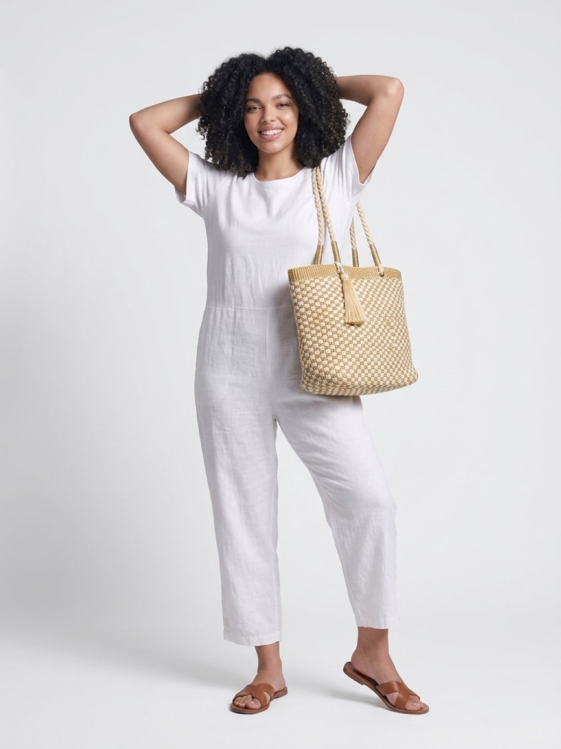 Smiling model wearing a white linen jumpsuit and leather sandals, carrying the elegant cream and gold metallic woven tote bag over her shoulder.