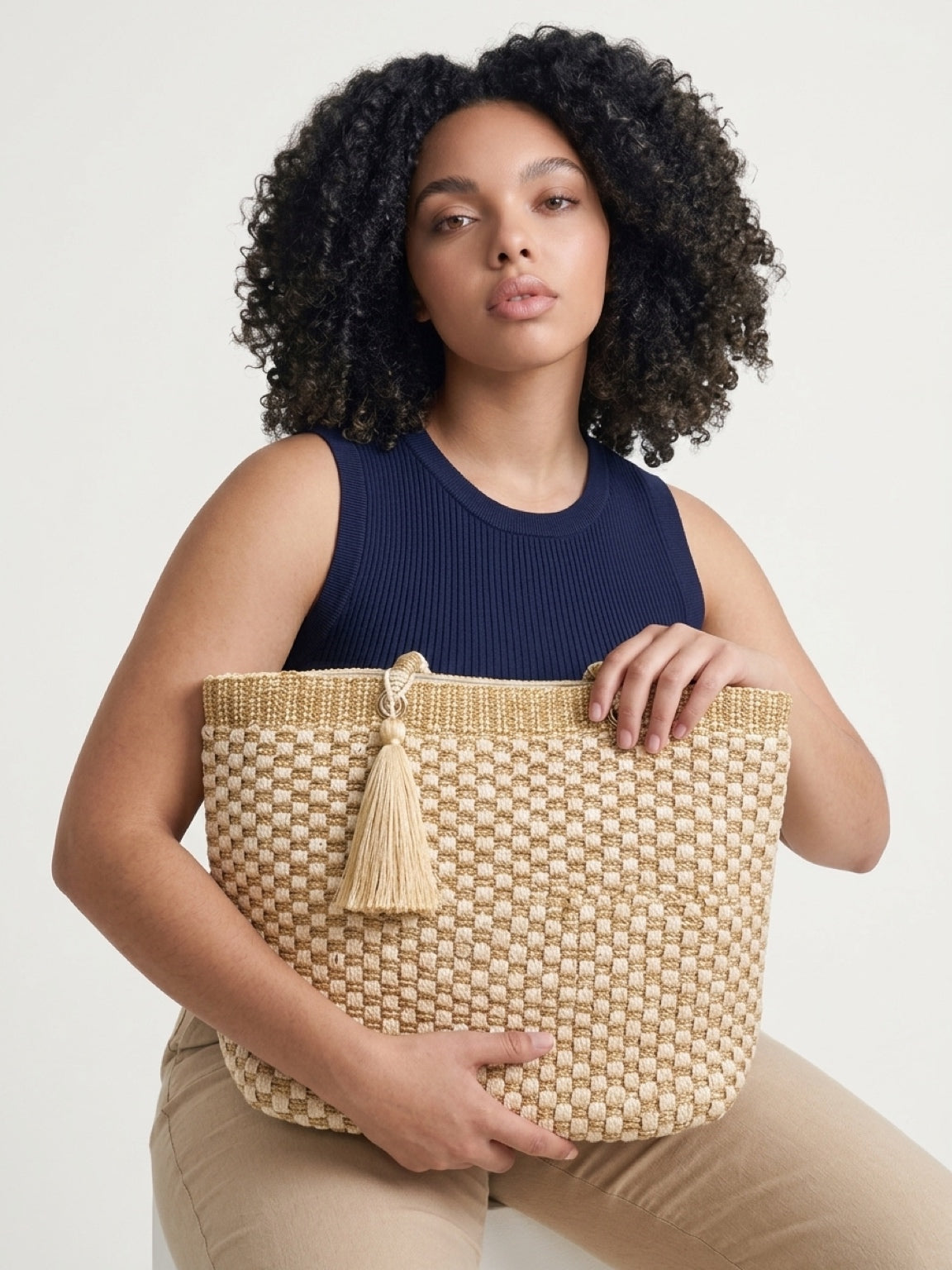 Close-up lifestyle shot of a model holding the cream and gold woven tote bag across her lap, highlighting the textured checkerboard pattern, top metallic border, and matching statement tassel.