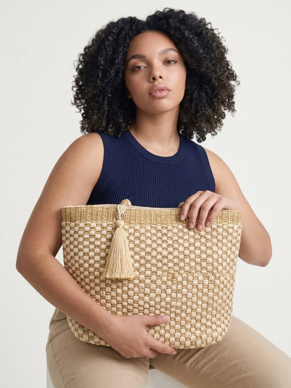 Close-up lifestyle shot of a model holding the cream and gold woven tote bag across her lap, highlighting the textured checkerboard pattern, top metallic border, and matching statement tassel.