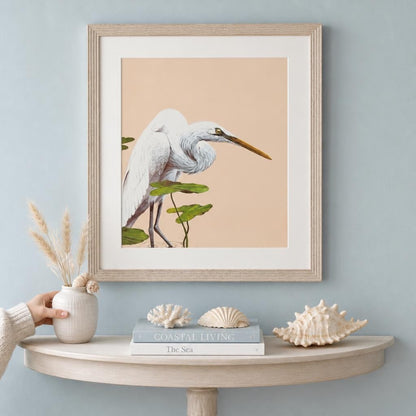 A framed vintage 1980 Great Egret wildlife art print styled in a coastal maximalist living room. The print is showcased in a light beige wood frame with a wide white mat board, hanging on a dusty blue wall above a white demi-lune console table. The table is decorated with seashell specimens, a white coral piece, decorative books titled "Coastal Living" and "The Sea", and a ceramic bud vase filled with dried pampas grass.