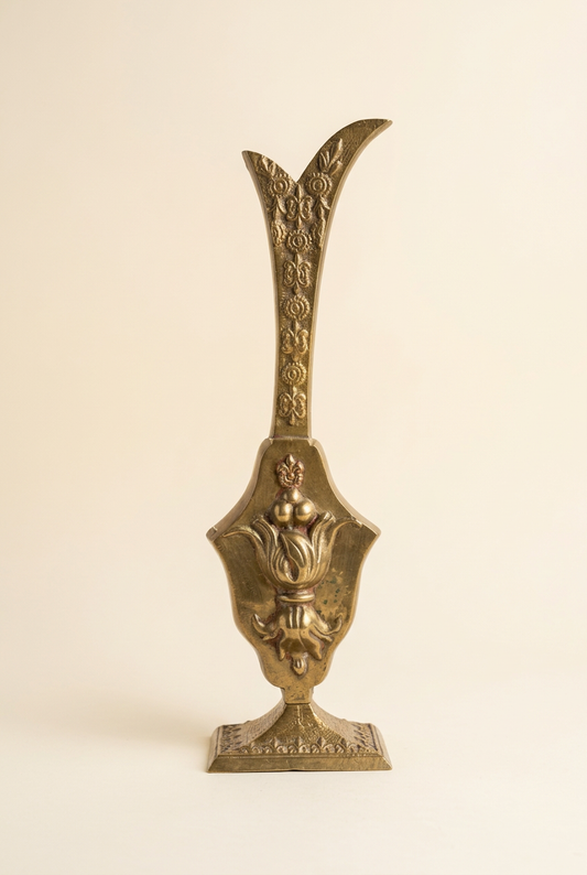  Ornate aged brass vessel with a tall flared neck, raised floral motifs, and a rectangular pedestal base, shown against a warm ivory background.