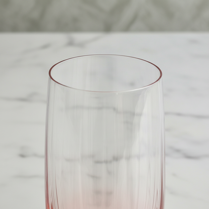 A close-up of a clear ribbed glass tumbler with a delicate pink gradient at the rim and base, set against a white marble background.
