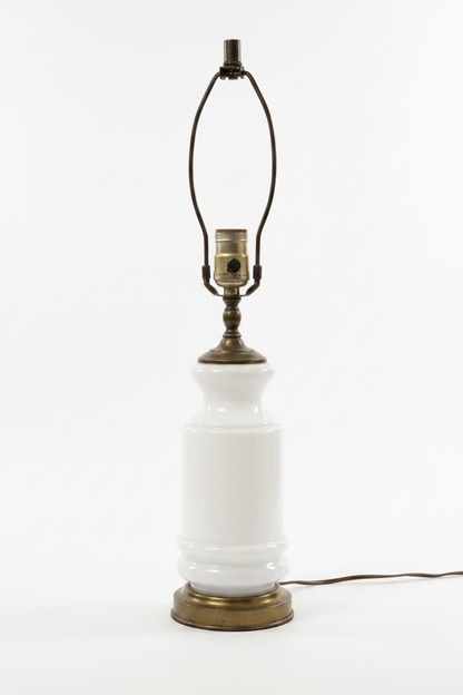 Vintage 1960s opaque white milk glass table lamp base shown without shade, stepped urn-to-cylinder profile with raised ring detail at lower body, antiqued brass shoulder cap and large circular brass plinth base with aged patina, brass harp and E26 turn-knob socket visible, brown cord, white background


