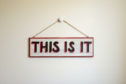 Hand-painted wooden sign reading THIS IS IT hanging on a neutral wall with natural jute rope, featuring red 3D shadow typography on a distressed white background.